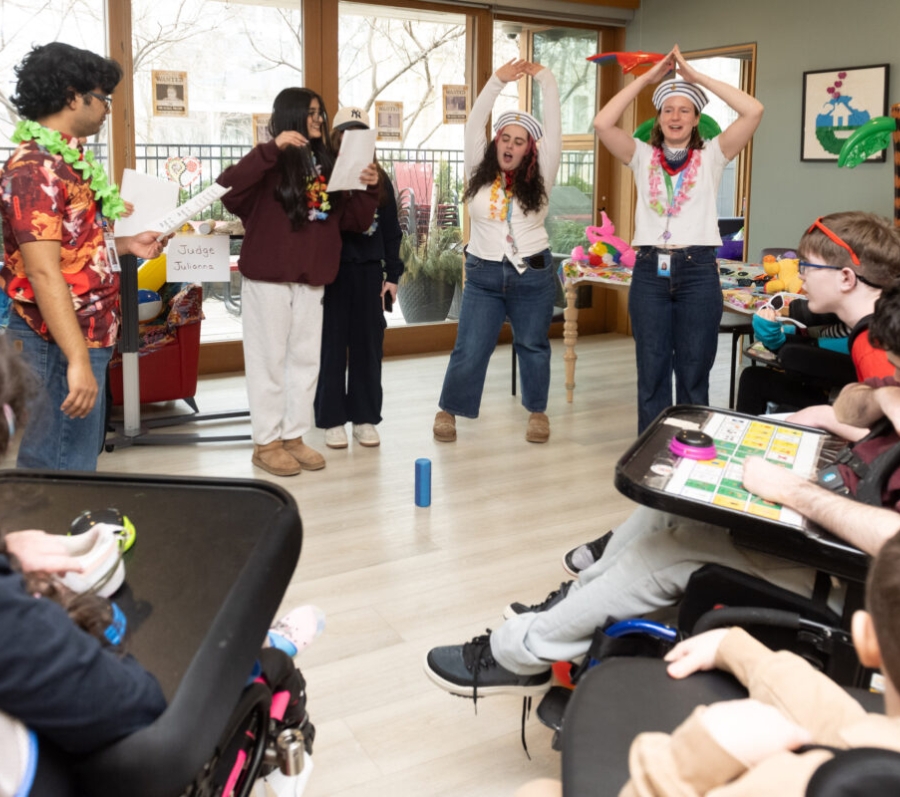 Group activity in a bright common room where several people stand in a circle participating in a game, while others sit nearby in wheelchairs and at tables, with large windows, decorations, and activity materials visible around the room.