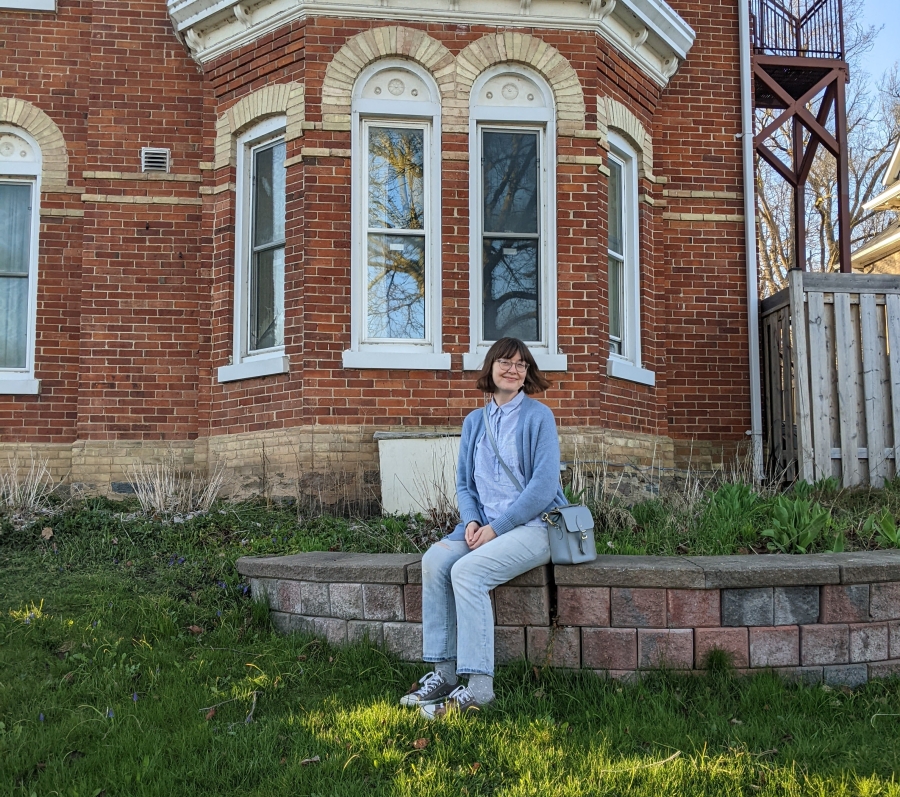 Person sitting on a low stone ledge in front of a red brick building with bay windows, surrounded by grass.