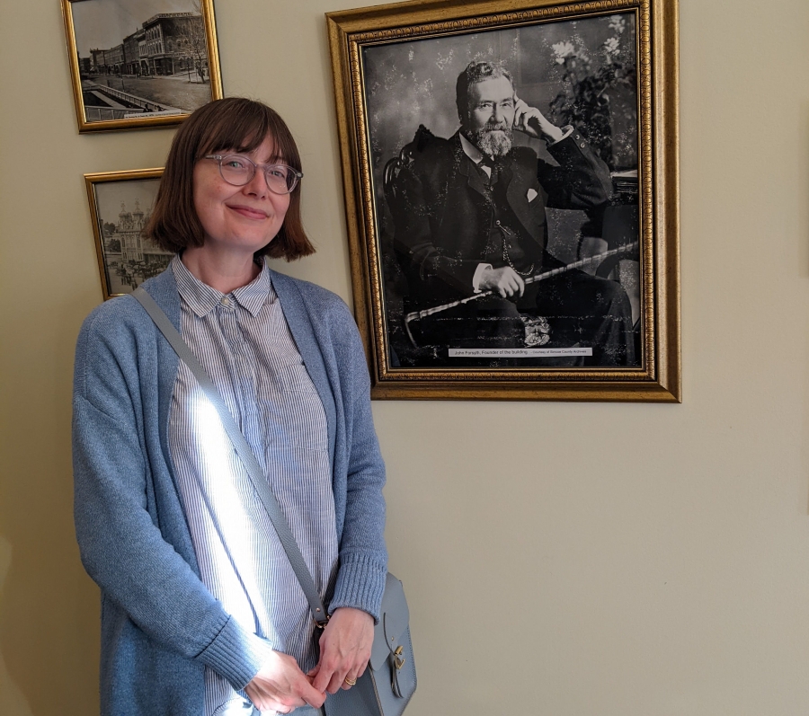 Person standing indoors beside framed photographs, smiling at the camera.