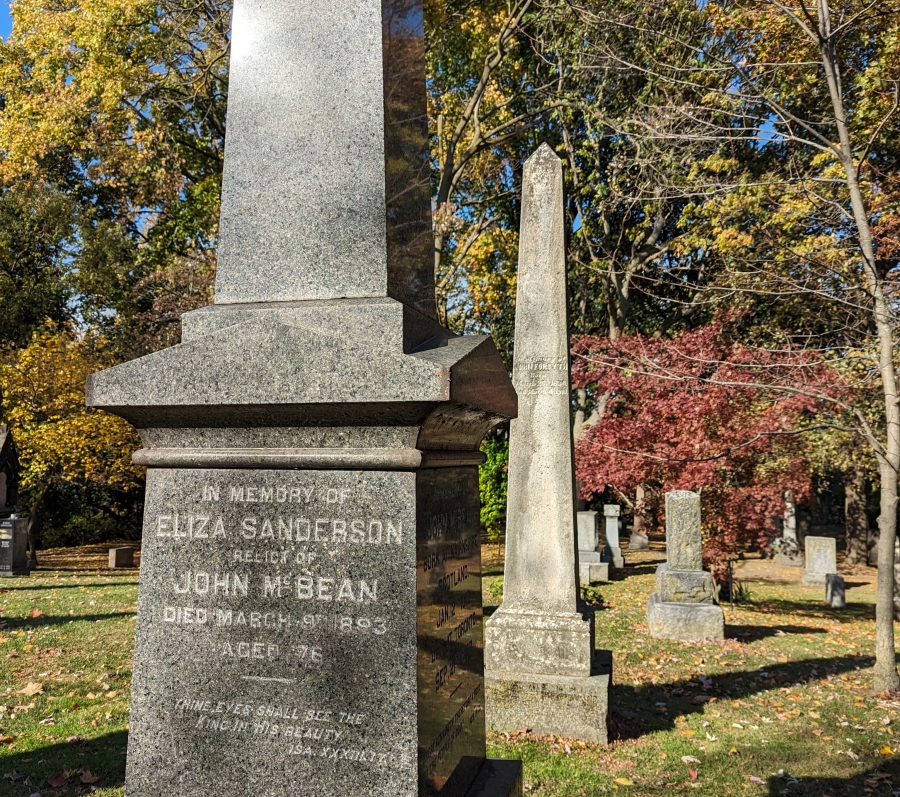 Tall stone monuments in a cemetery during autumn, with trees and fallen leaves in the background.