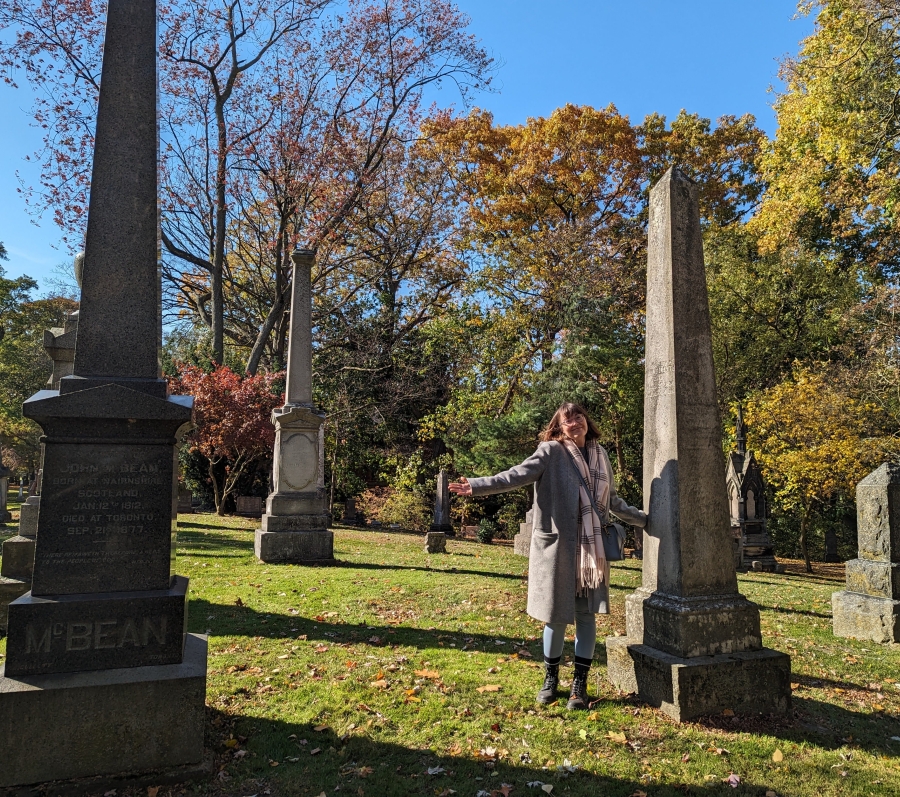 Person standing among tall gravestones in a cemetery, gesturing with arms out, with autumn trees in the background.