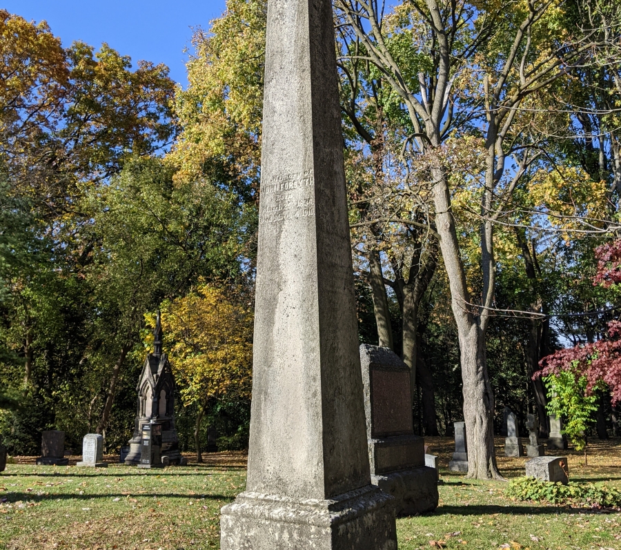 Tall stone obelisk monument in a cemetery, surrounded by trees with autumn foliage.