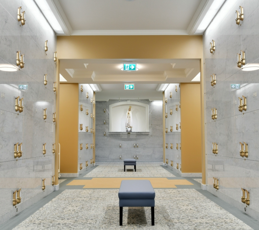 An elegant indoor mausoleum hallway with polished light‑gray marble walls featuring rows of bronze crypt handles. The space has warm beige accent walls, overhead lighting, and a terrazzo-style floor. A small padded bench sits in the center, and at the far end is a niche holding a religious statue on a marble pedestal.