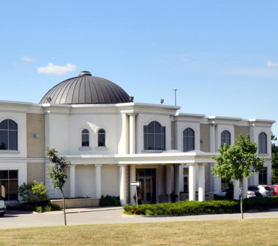 A two‑storey beige and white building with a large dark domed roof centered above the entrance. The front features tall arched windows and columns leading to a covered entryway. Several cars are parked along the driveway, with small trees and shrubs in front of the building under a clear blue sky.