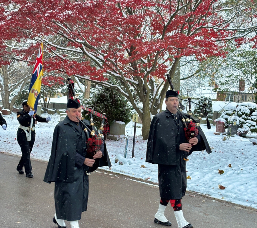 Bagpipers in dark uniforms marching on a snowy path with a flag bearer, red autumn tree in background.