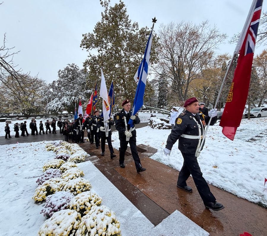 Uniformed honor guard marching on a snowy path carrying multiple flags, with flower arrangements in the foreground.