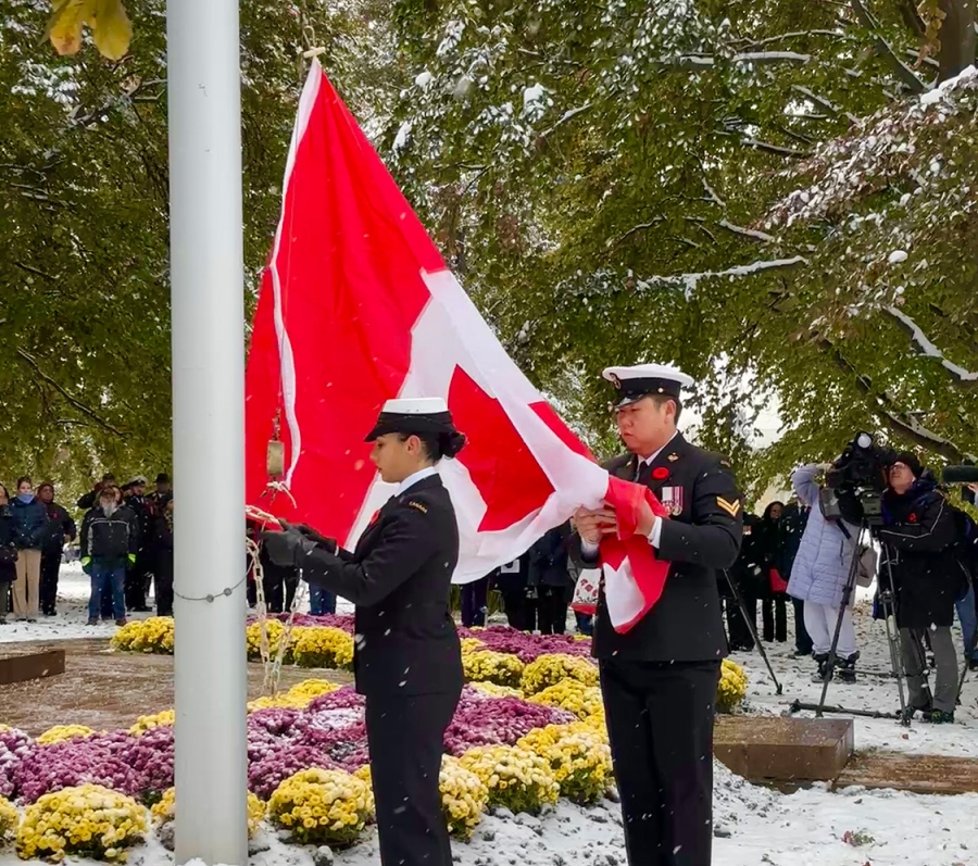 Two uniformed service members raise a Canadian flag on a snow-covered flagpole during a ceremony, with onlookers gathered among winter trees and flowers.