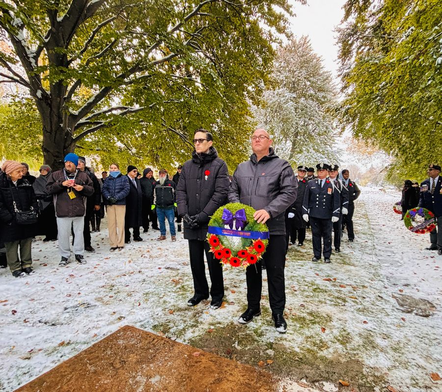 Two people holding a wreath with red poppies during a snowy remembrance ceremony, with others standing behind.