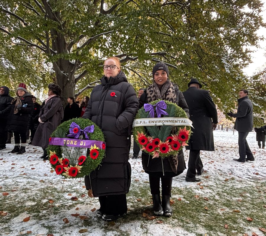 Two people holding wreaths with red poppies and purple ribbons during a snowy remembrance ceremony, with others gathered behind.