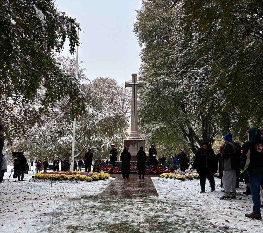 Snowy remembrance ceremony at a war memorial with a large stone cross, wreaths arranged at its base, and people gathered around.