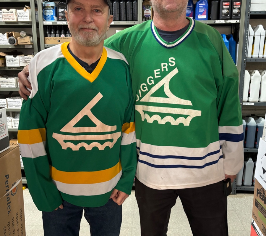 Two men standing side by side in a store aisle, wearing green hockey jerseys, with shelves of automotive supplies behind them.