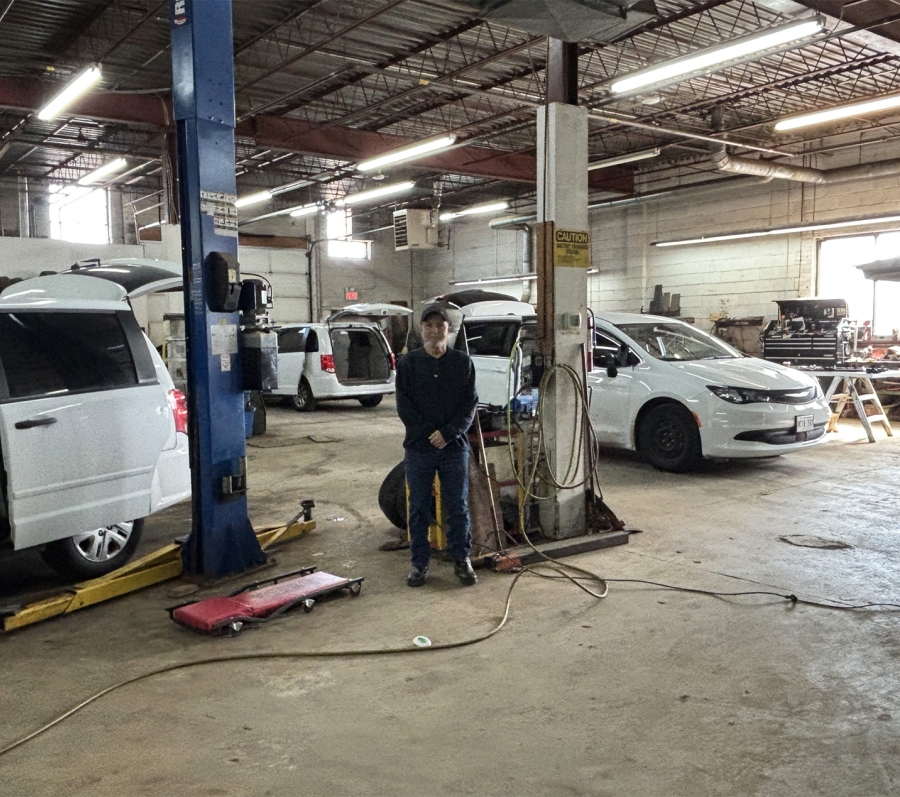Man standing inside an auto repair shop with several vans on lifts and open hoods, surrounded by tools and service equipment.
