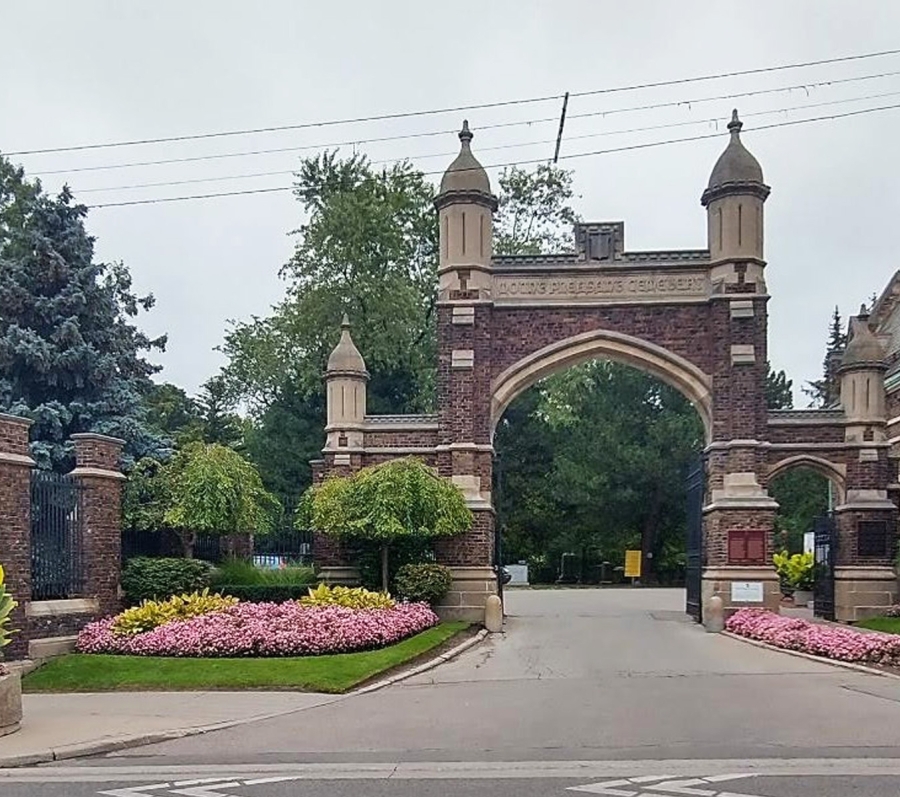 Main entrance to Mount Pleasant Cemetery, featuring a large stone and brick archway with decorative pillars, iron gates, and landscaped flower beds lining the roadway.