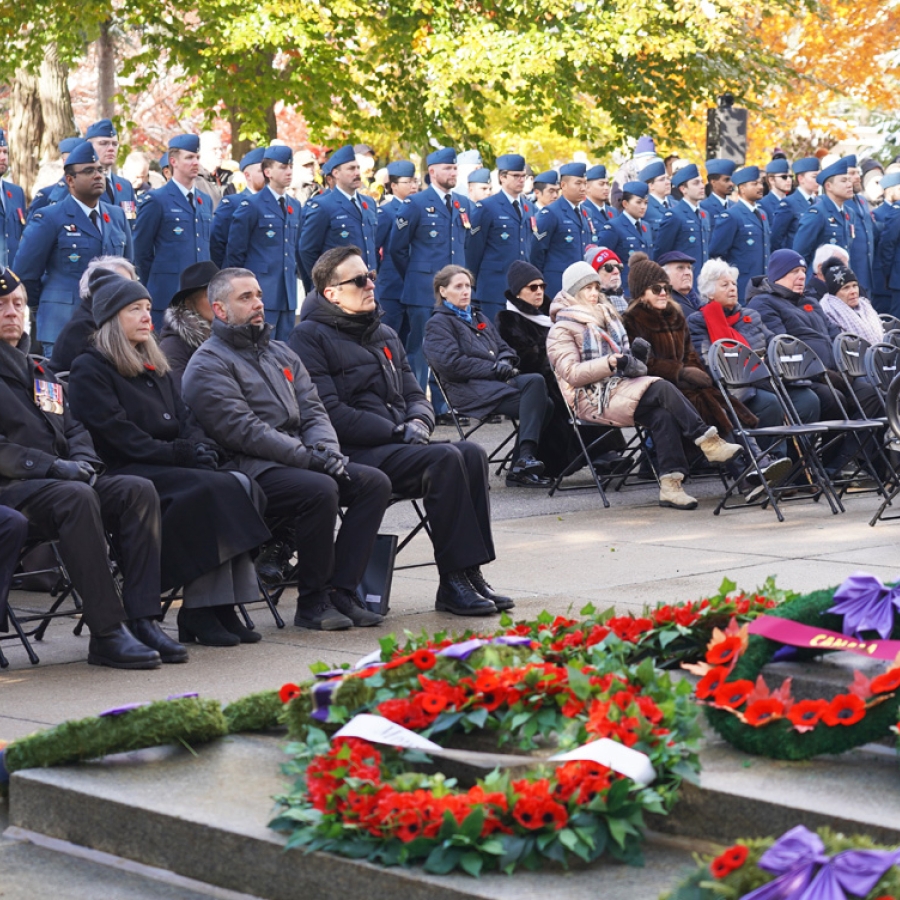 People seated at an outdoor memorial ceremony with wreaths displayed in the foreground.