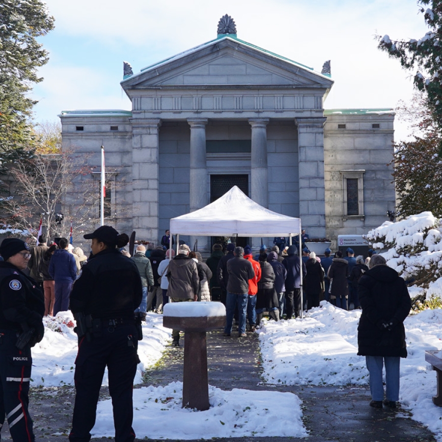 A crowd gathers outside a stone building during a winter memorial event, with snow lining the walkway.