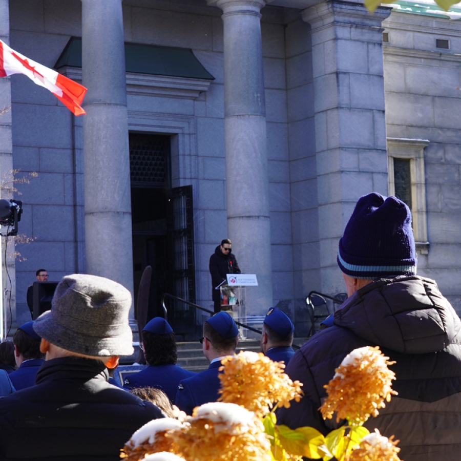 A speaker stands at a podium outside a stone building during a memorial ceremony, with attendees gathered in the foreground and a Canadian flag flying nearby.