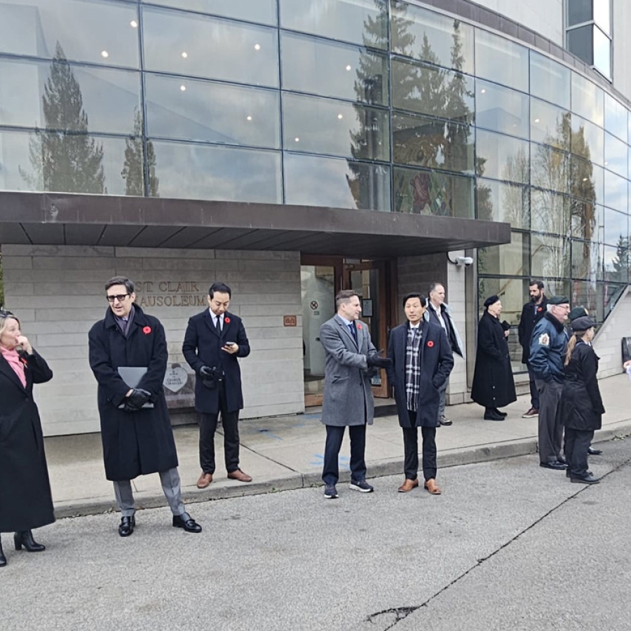 People stand outside a modern building during a remembrance gathering, some wearing coats and poppies along a sidewalk.