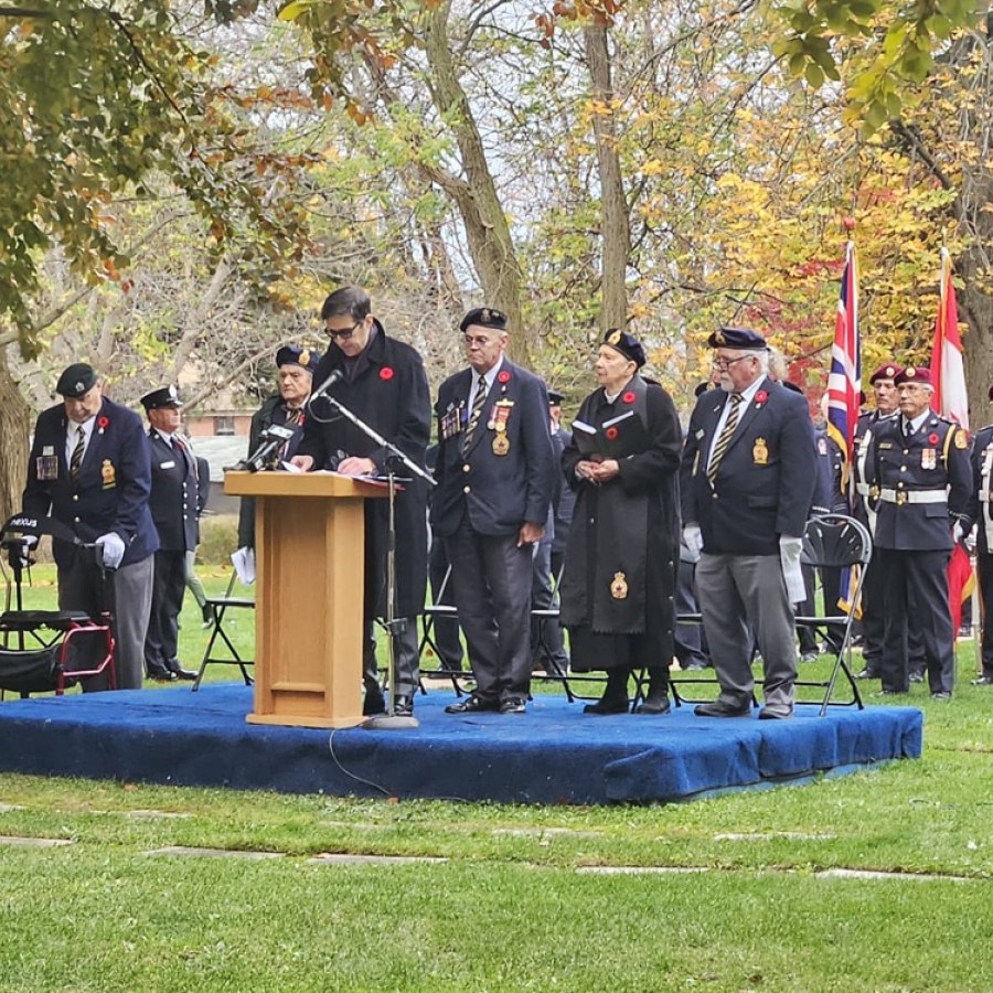 Veterans stand on a small outdoor stage near a podium during a remembrance ceremony, with flags displayed behind them.