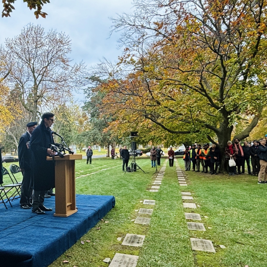 A speaker stands at a podium on an outdoor stage during a remembrance ceremony, with attendees gathered among trees and memorial stones.