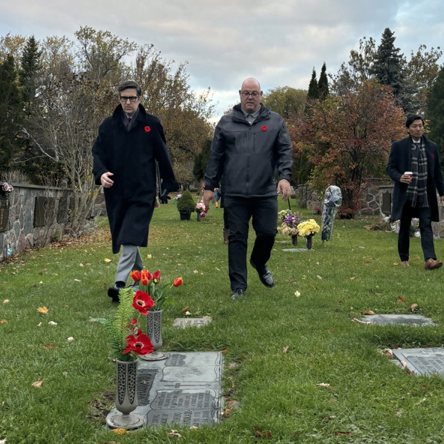 Three people walk through a cemetery during a remembrance visit, with headstones, flowers, and autumn trees visible around them.
