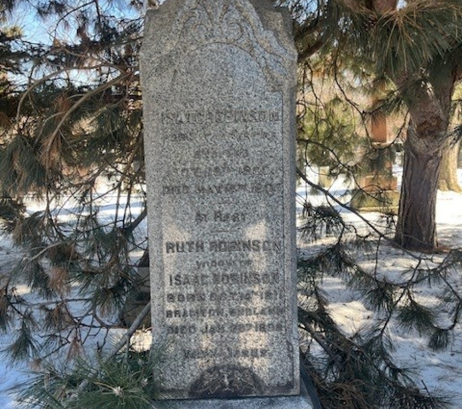 Weathered gray stone grave marker with an arched top, partially obscured by pine tree branches, standing in a snowy cemetery; the engraved inscription is faint and difficult to read.