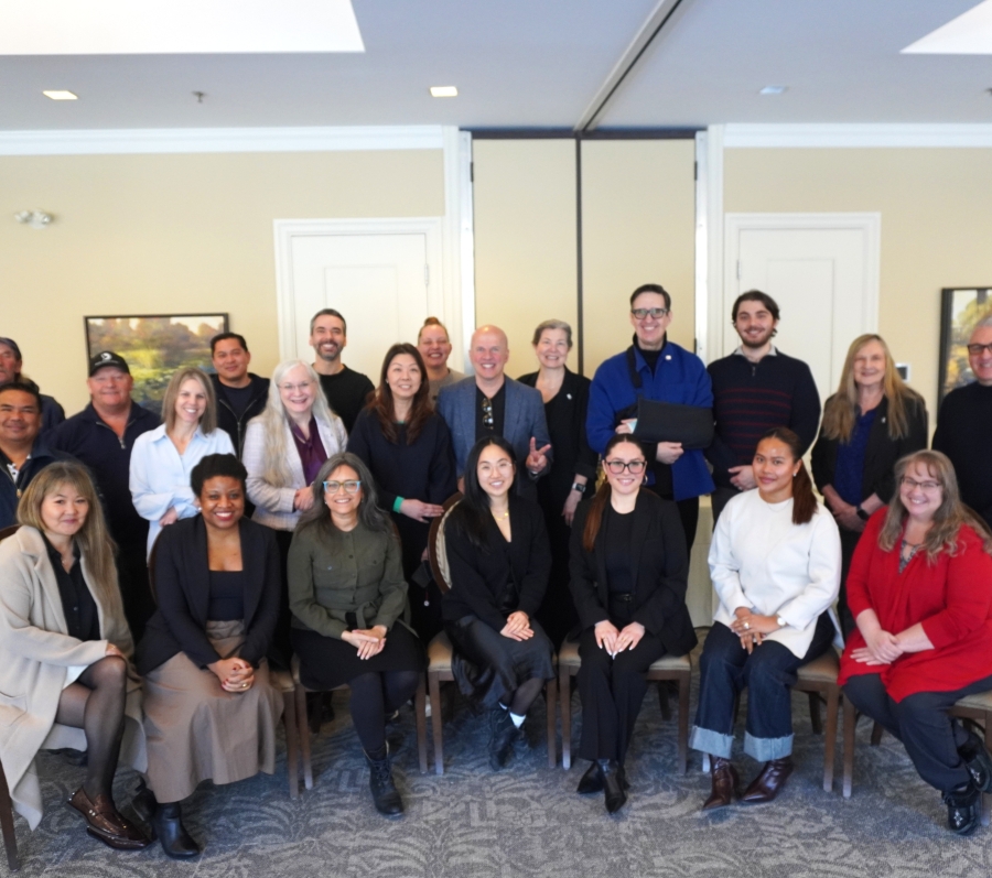 Group photo of about twenty people seated and standing in a conference room with beige walls, framed artwork, patterned carpet, and a folding partition behind them.