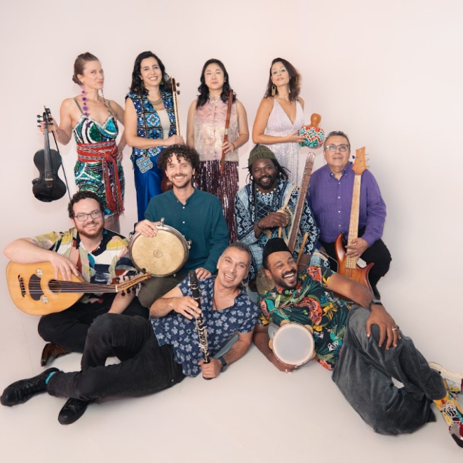 Group of musicians posing together with various instruments, including drums, violin, and guitar, against a plain background.