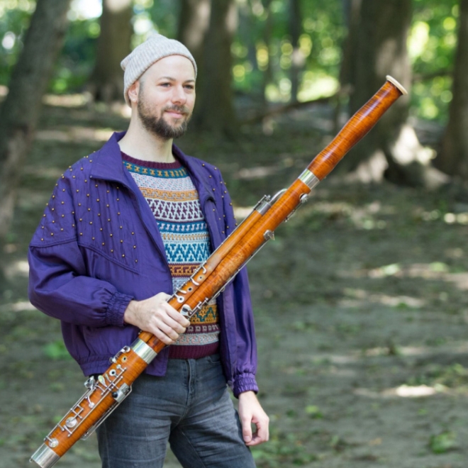 Person standing outdoors in a wooded area holding a large bassoon, wearing a jacket and knit hat.