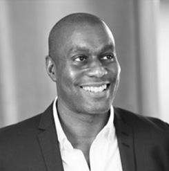Professional headshot of a person in a dark blazer and open-collar shirt, smiling against a blurred background.