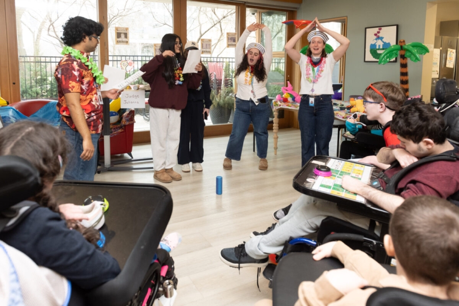Group activity in a bright common room where several people stand in a circle participating in a game, while others sit nearby in wheelchairs and at tables, with large windows, decorations, and activity materials visible around the room.