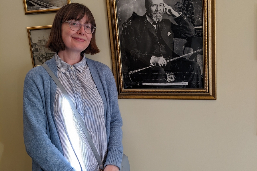 Person standing indoors beside framed photographs, smiling at the camera.