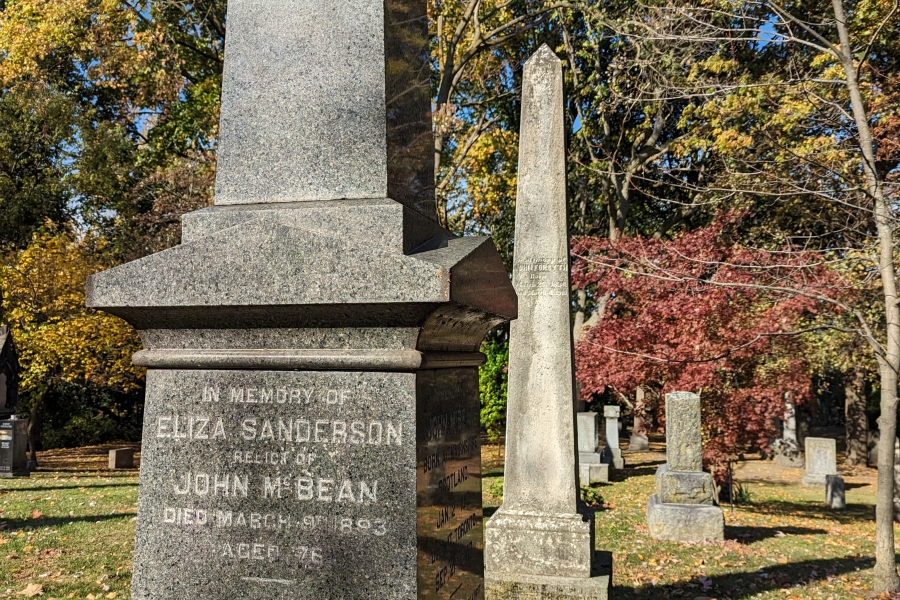 Tall stone monuments in a cemetery during autumn, with trees and fallen leaves in the background.
