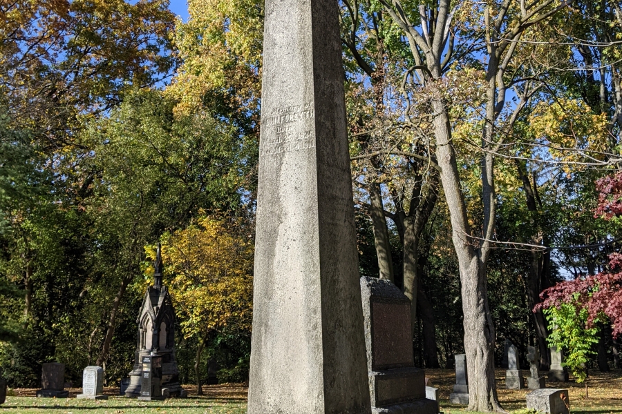 Tall stone obelisk monument in a cemetery, surrounded by trees with autumn foliage.