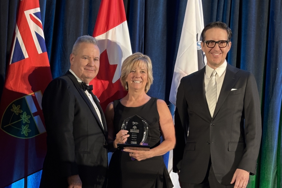Three people in formal attire pose on stage with flags behind them as a woman holds an award.