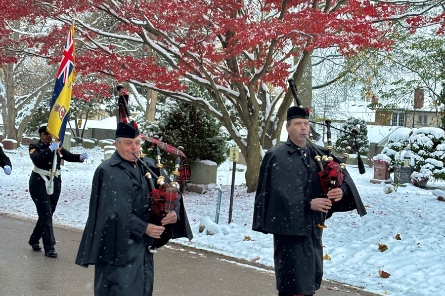 Bagpipers in dark uniforms marching on a snowy path with a flag bearer, red autumn tree in background.