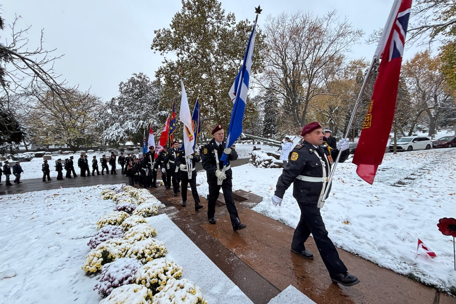 Uniformed honor guard marching on a snowy path carrying multiple flags, with flower arrangements in the foreground.