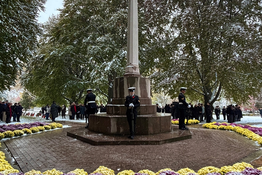 A tall stone cross monument surrounded by snow-dusted flowers, with uniformed guards standing watch and people gathered in the background.