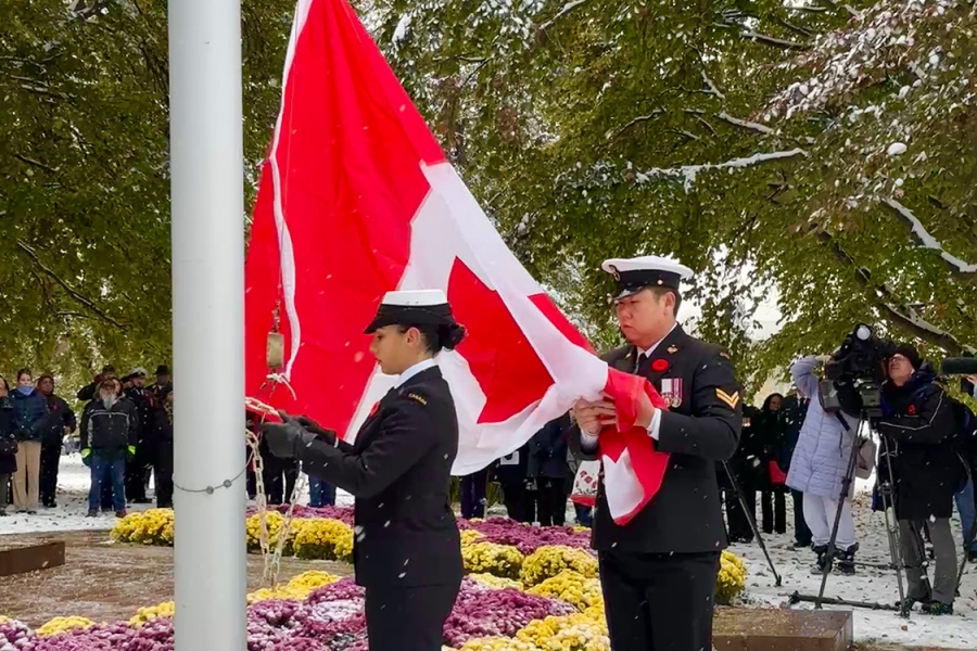Two uniformed service members raise a Canadian flag on a snow-covered flagpole during a ceremony, with onlookers gathered among winter trees and flowers.