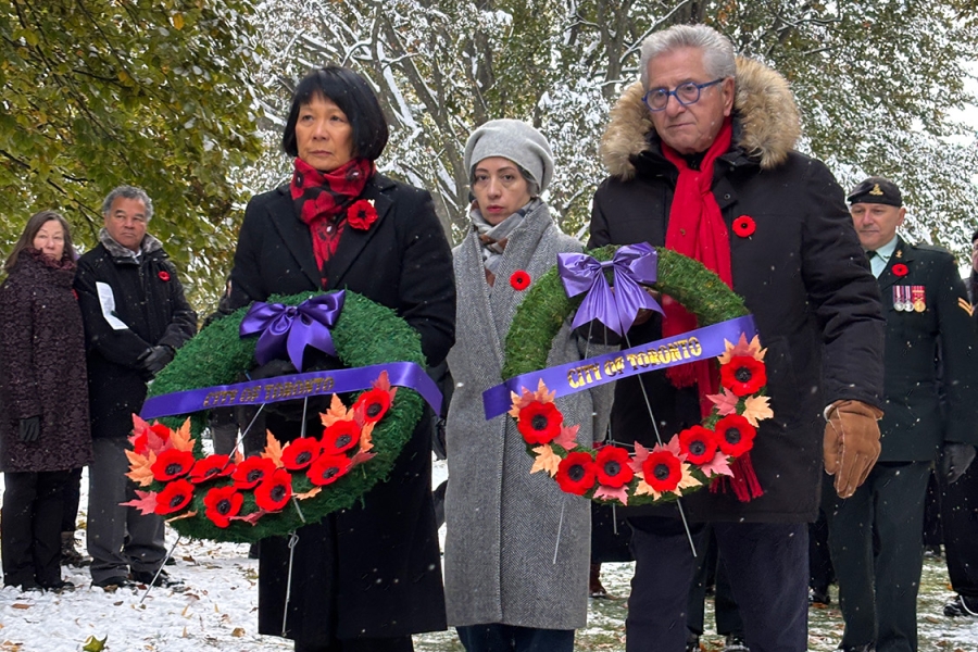 People at an outdoor remembrance ceremony holding wreaths with red poppies and purple ribbons on a snowy day.