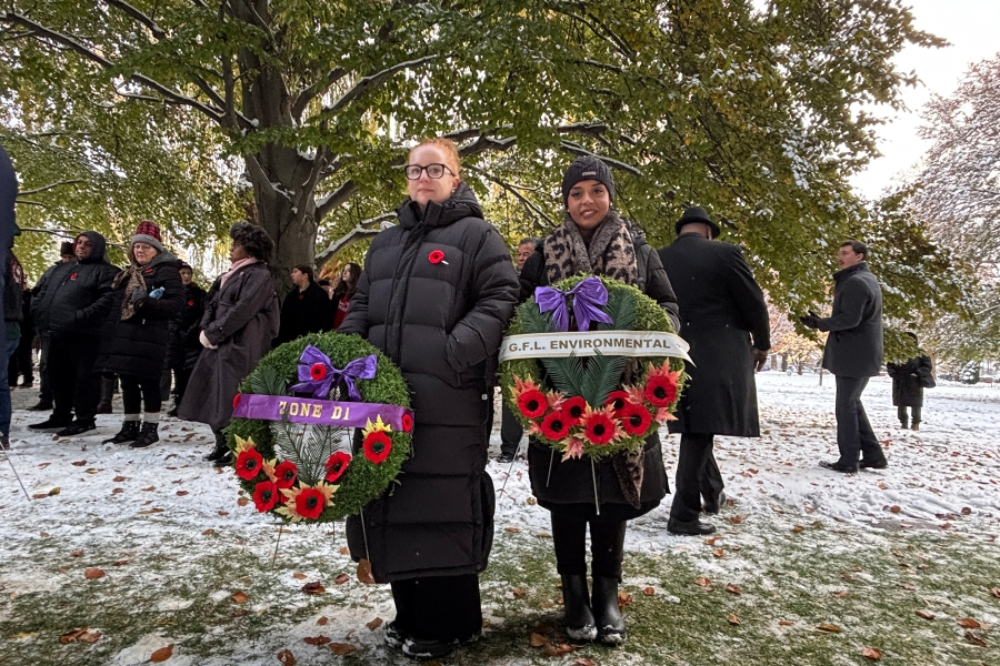 Two people holding wreaths with red poppies and purple ribbons during a snowy remembrance ceremony, with others gathered behind.
