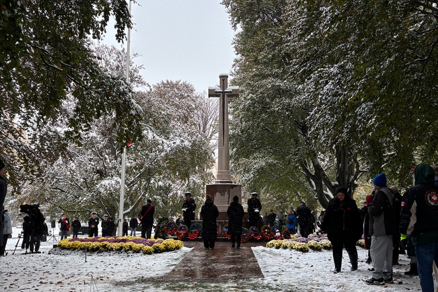 Snowy remembrance ceremony at a war memorial with a large stone cross, wreaths arranged at its base, and people gathered around.