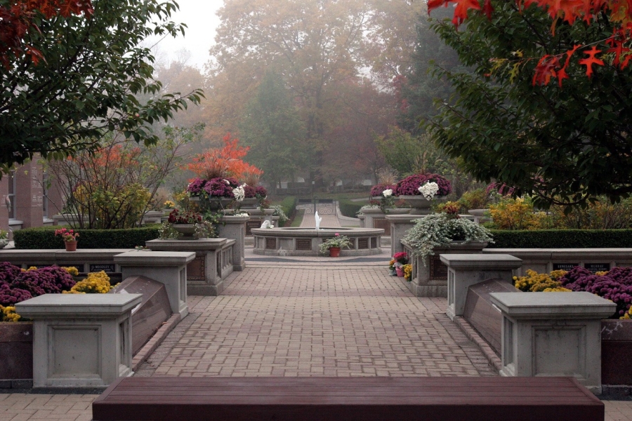 Cemetery garden path with flower-filled planters and central fountain in autumn mist.