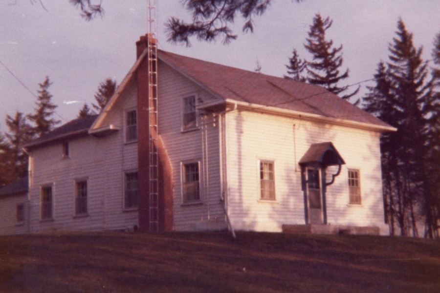 Small white rural building with a sloped roof and chimney, surrounded by trees.