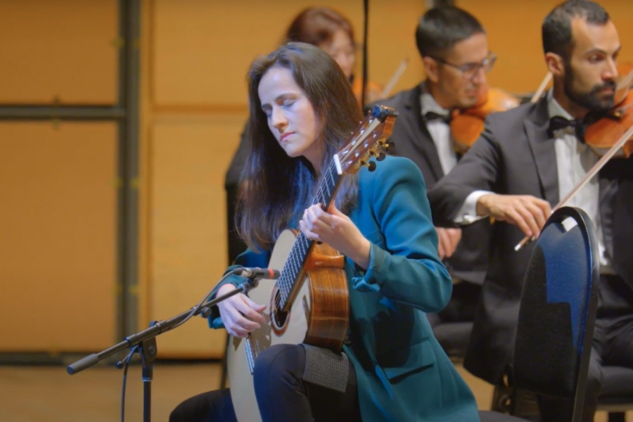 Classical guitarist performing on stage with a microphone, accompanied by an orchestra in the background.