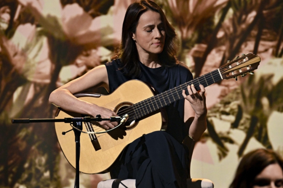 Person playing a classical guitar on stage with a microphone, against a blurred backdrop.