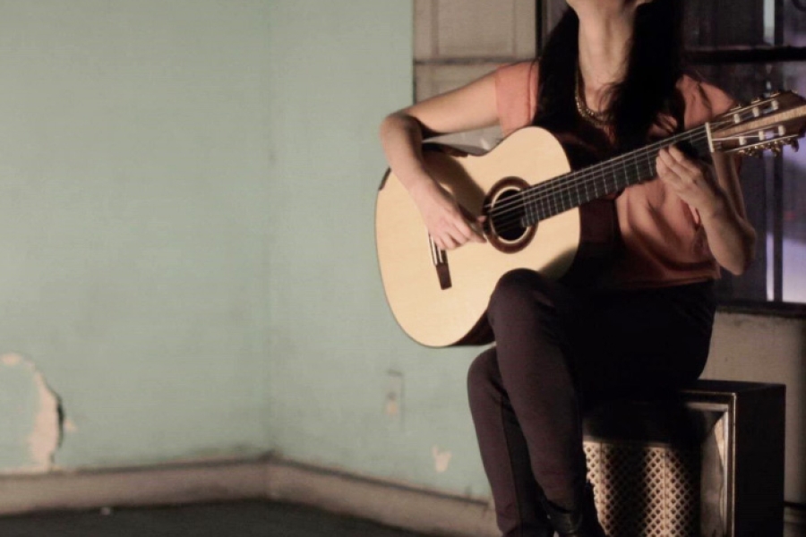 Person seated indoors playing a classical guitar, lit by warm light against a simple wall background.