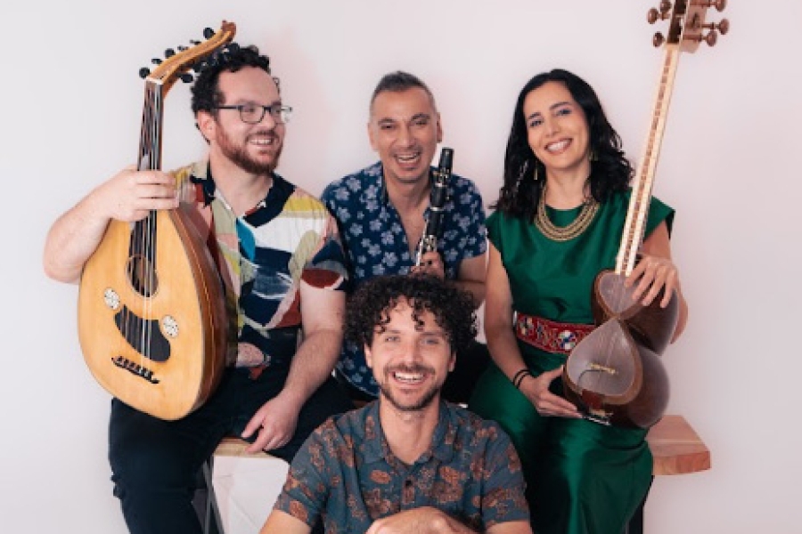 Four musicians posing together with string and percussion instruments against a plain background.