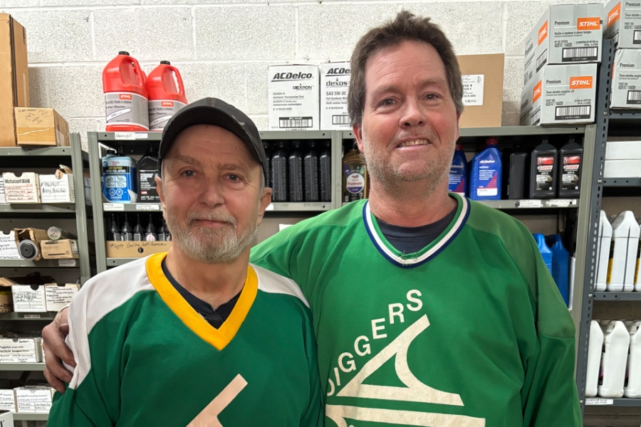 Two men standing side by side in a store aisle, wearing green hockey jerseys, with shelves of automotive supplies behind them.