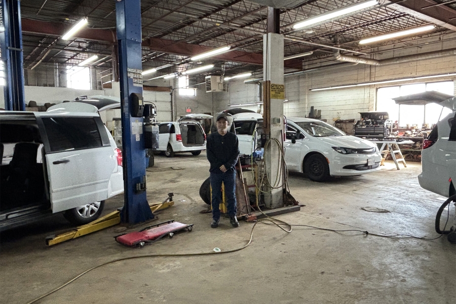 Man standing inside an auto repair shop with several vans on lifts and open hoods, surrounded by tools and service equipment.
