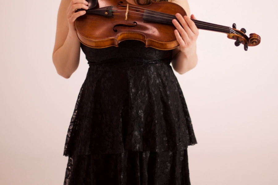 A person in a black dress holding a violin, looking down at the instrument against a plain background.